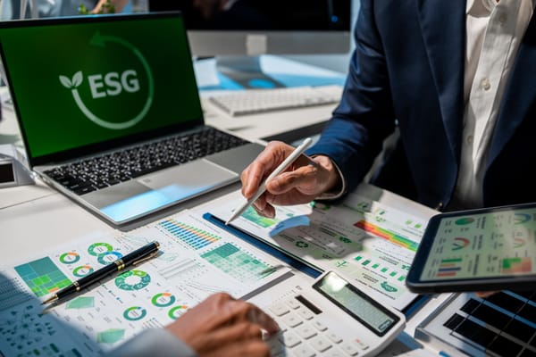 A person marking up printed sustainability charts with a stylus, beside a laptop showing an ESG logo, a tablet and a calculator